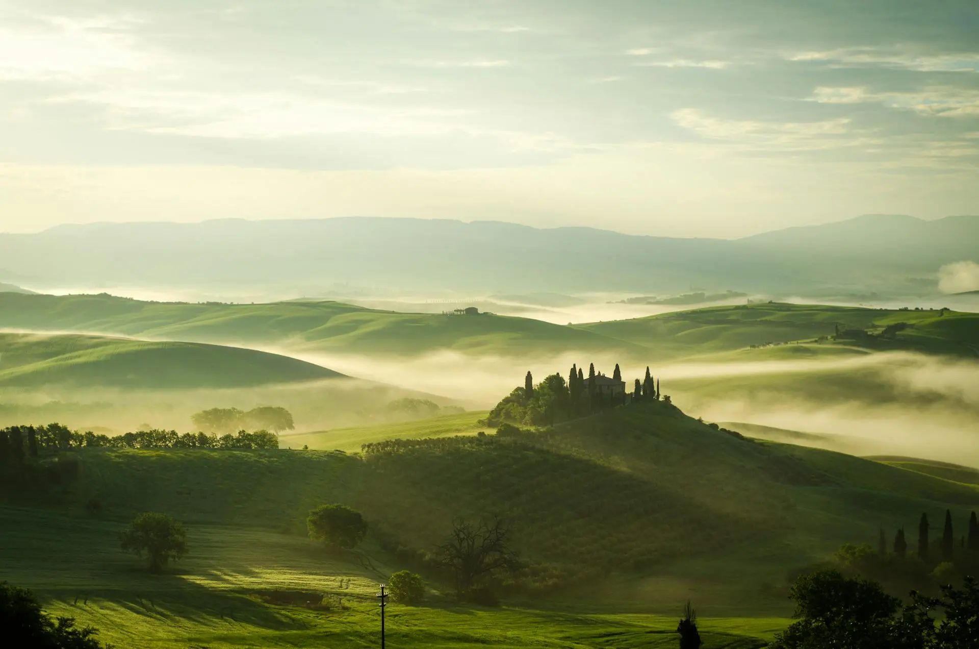 Misty Tuscan landscape with rolling hills: A scenic view of green hills partially obscured by mist, featuring a hilltop village in the distance. Ideal for promoting scenic tours and landscape photography in Tuscany.